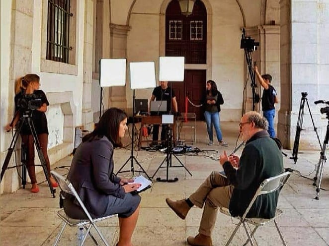 Entrevista montada num pátio de arcadas, duas pessoas sentadas frente a frente, com luzes e câmaras em tripé à volta.